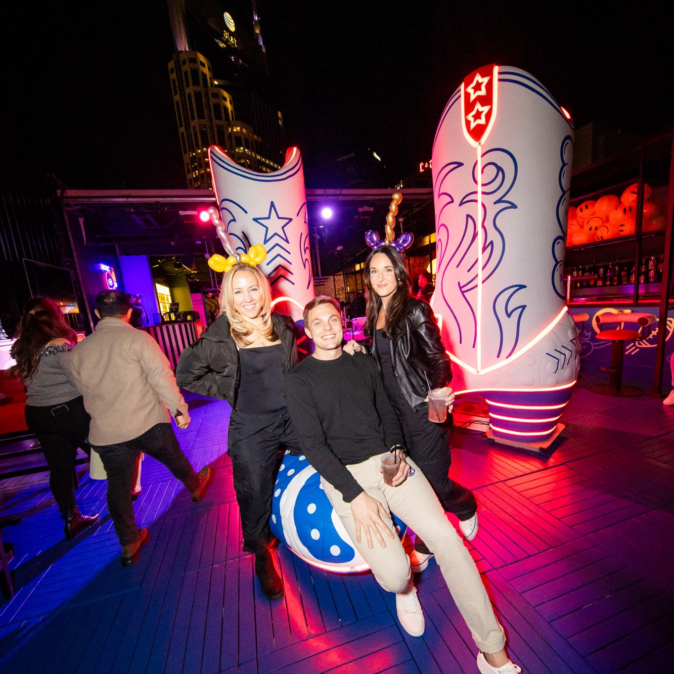 Two women and a man enjoying an evening on the rooftop of Play Playground at Nashville Live! Oversized boots and skyline are in the background.