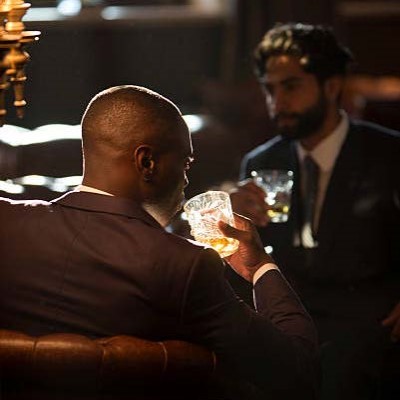 Two men in suits drinking cocktails and sitting in leather club chairs in a dark bar lounge.